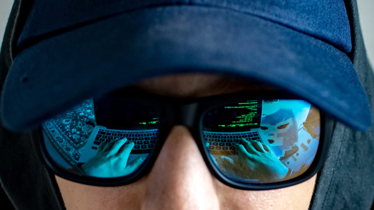 In der Sonnenbrille eines jungen Mannes mit schwarzem Hoodie und Basecap spiegelt sich ein Laptop mit grüner Schrift auf schwarzem Hintergrund. Man sieht seine Hände auf der Tastatur und einen gruseligen Sticker daneben.