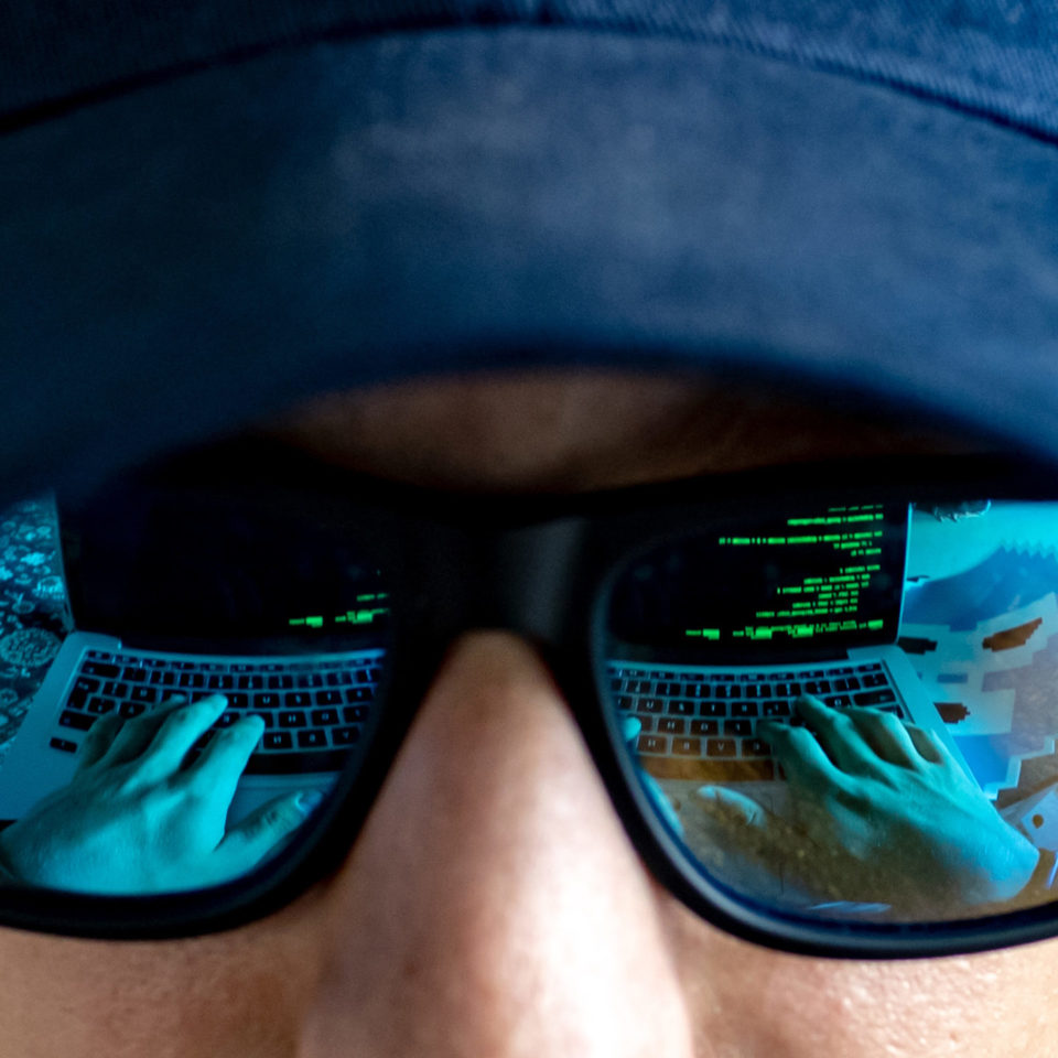 In der Sonnenbrille eines jungen Mannes mit schwarzem Hoodie und Basecap spiegelt sich ein Laptop mit grüner Schrift auf schwarzem Hintergrund. Man sieht seine Hände auf der Tastatur und einen gruseligen Sticker daneben.